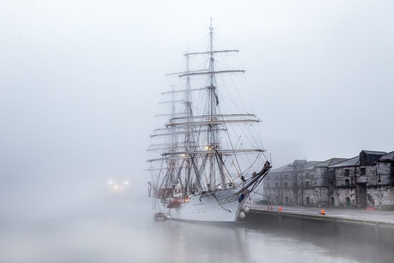 A full rigged sailing ship docked at a quay on a foggy morning.