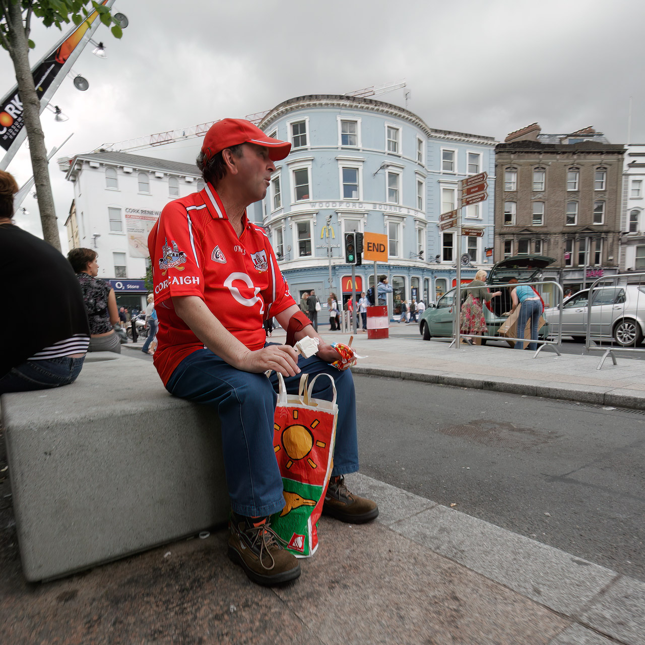 A man wearing a Cork jersey sits on a stone block by Daunt Square in Cork while he eats an ice lol.