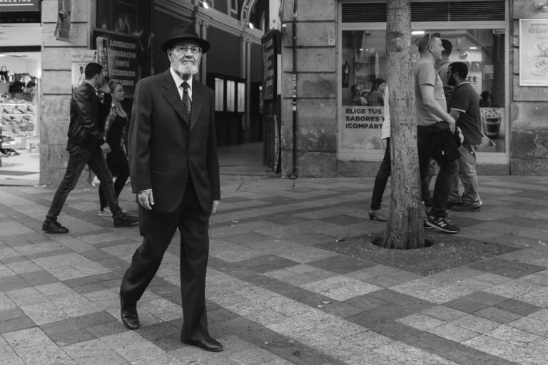 A black and white photo of a man dressed in a suit and hat walking on a pedestrianised street while other people in the background walk by.