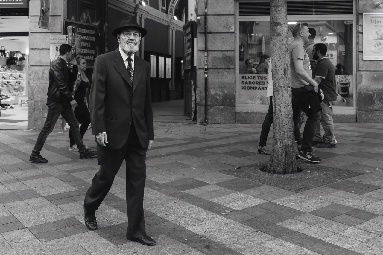 A black and white photo of a man dressed in a suit and hat walking on a pedestrianised street while other people in the background walk by.