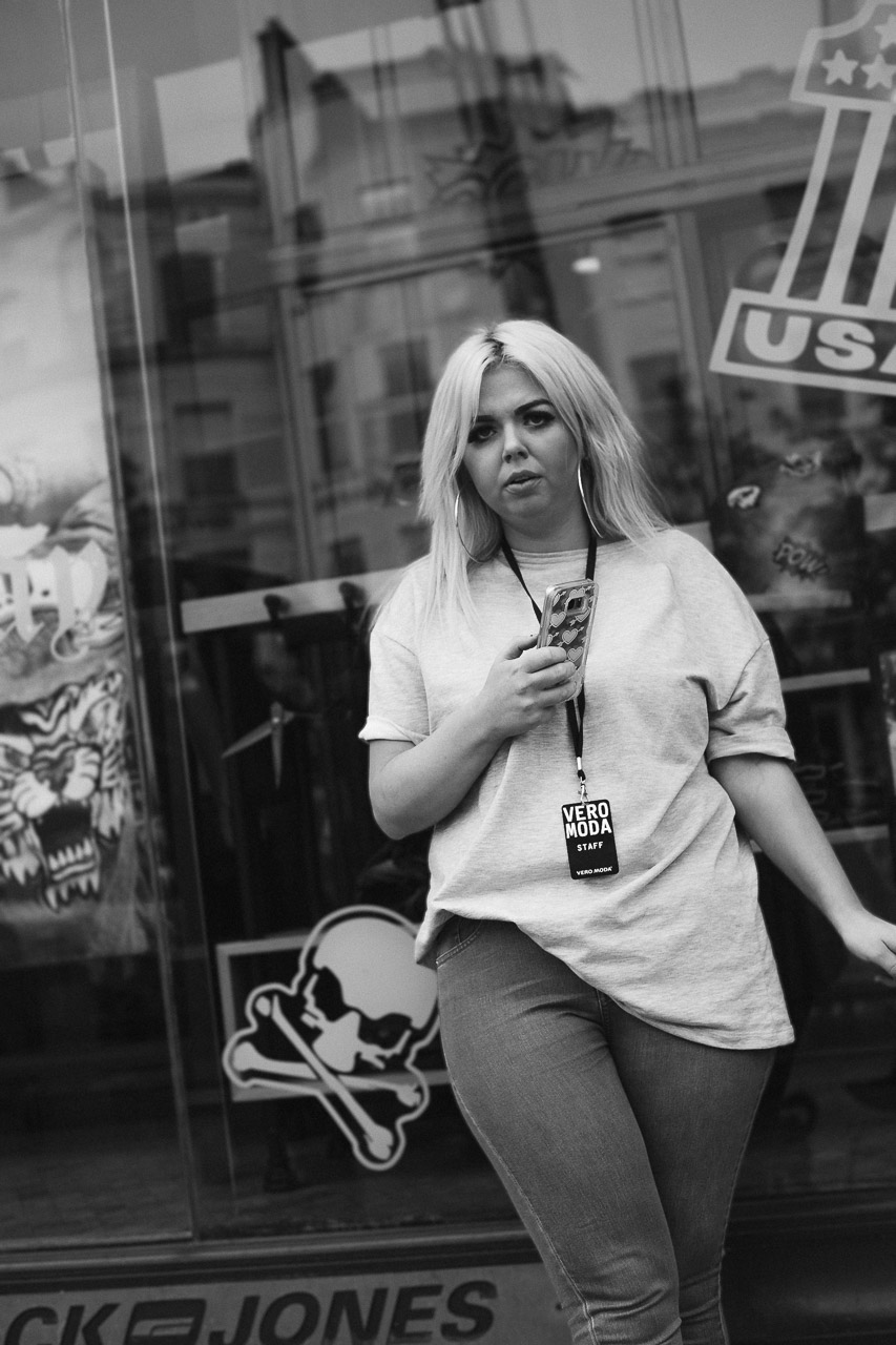 A woman leaning against a shop window stares at the camera. She has a mobile phone in her hand and a lanyard around her neck says, "Vero Moda staff"