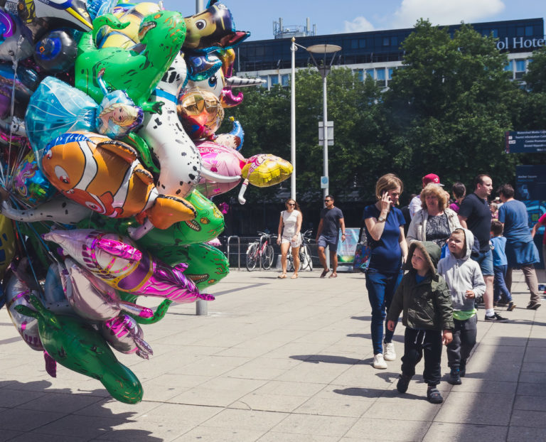 Kids walking along with some adults watch a large bunch of balloons for sale by a vendor who is just out of shot