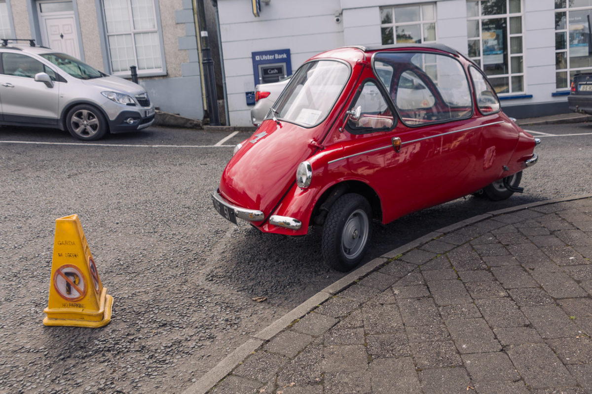 A small red car parked by a pavement with a "no parking" cone in front of it. It appears to be parked on a corner.