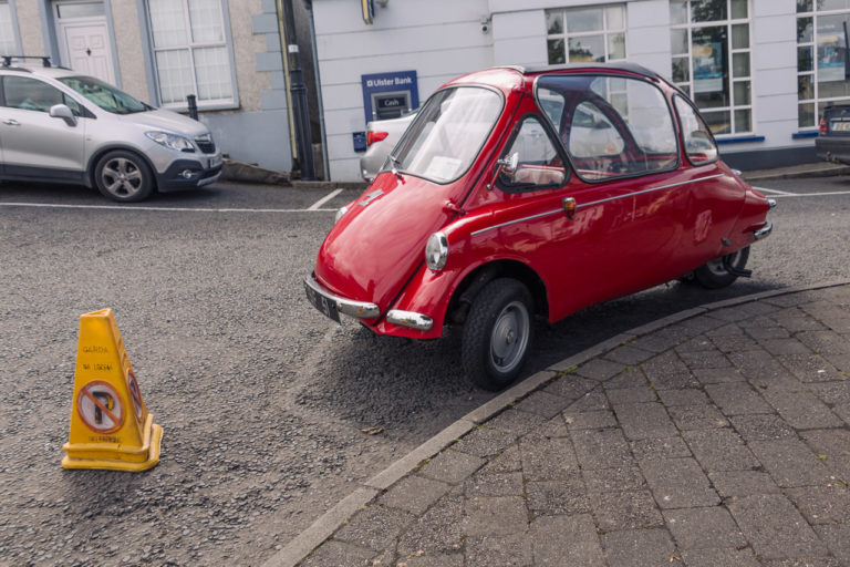 A small red car parked by a pavement with a "no parking" cone in front of it. It appears to be parked on a corner.