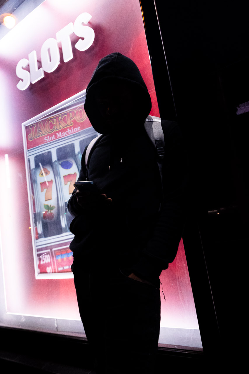 A young man checks his phone while leaning on a building. The word Slots appears in the window behind him.