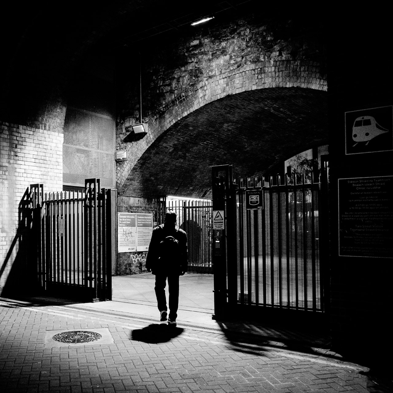 Black and white image of a shadowed man walking through gates towards a train station