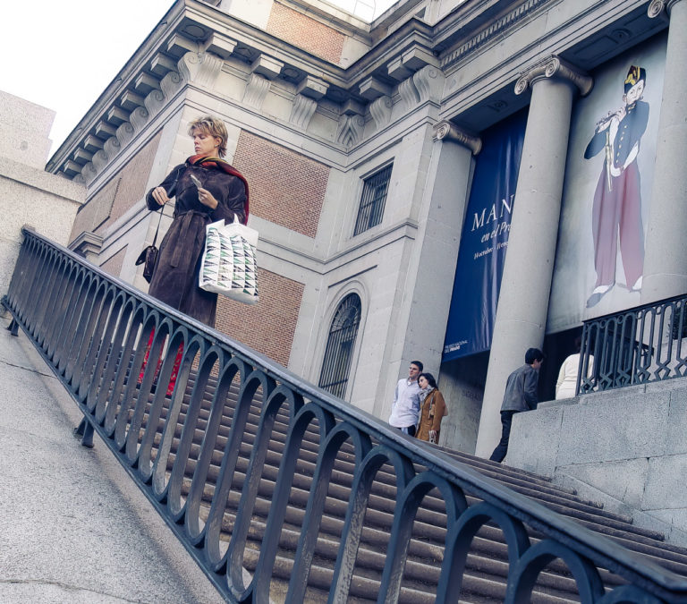 A woman adjusts her handbag on the steps of the Museo Nacional del Prado in Madrid. A couple have their photo taken in the background. Large signs show the text "MANE en el Prado" and some dates. Two people enter the museum.
