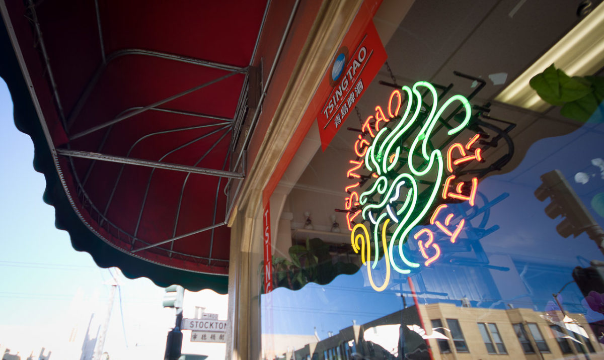 The window of a restaurant with an overhanging cover. The street is refected in the window and a sign for Tsingtao Beer floats in the window.