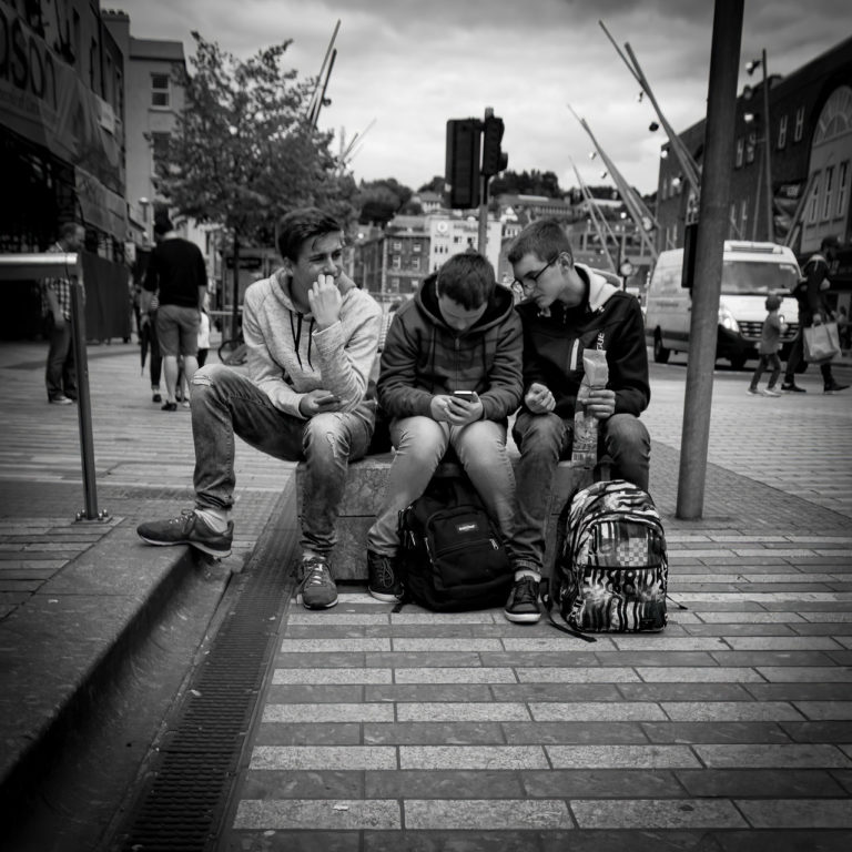 Teenage boys sit on a block on a street. One bites his nails looking worried, the middle guy is busy texting, and the last boy looks over his shoulder.