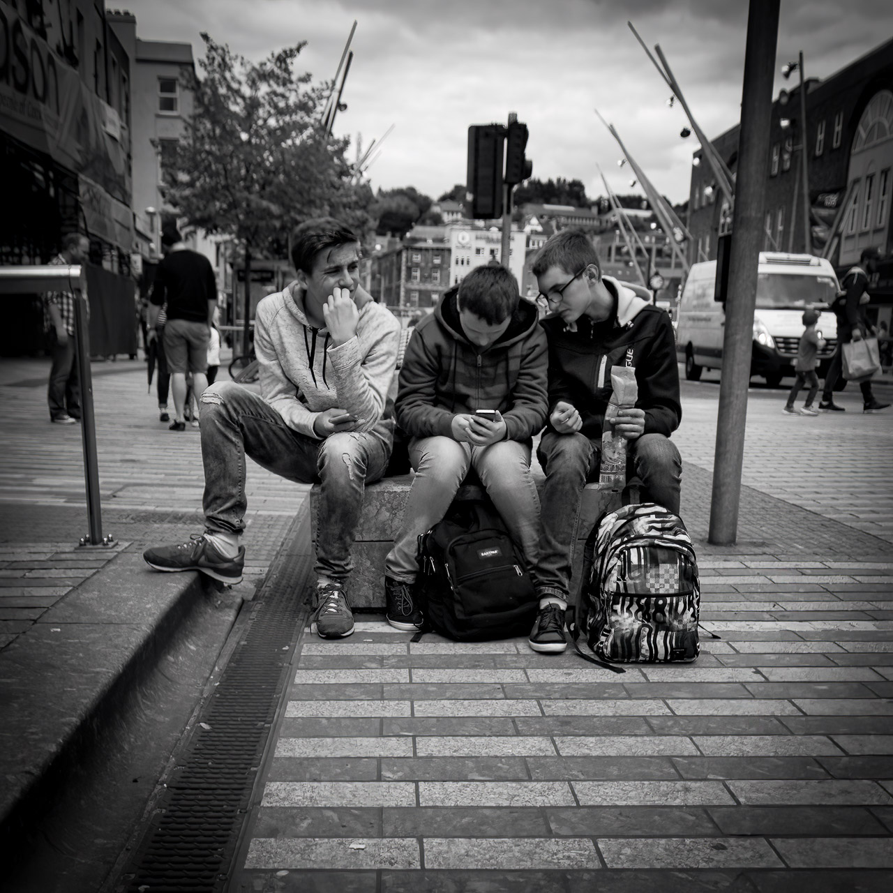 Teenage boys sit on a block on a street. One bites his nails looking worried, the middle guy is busy texting, and the last boy looks over his shoulder.