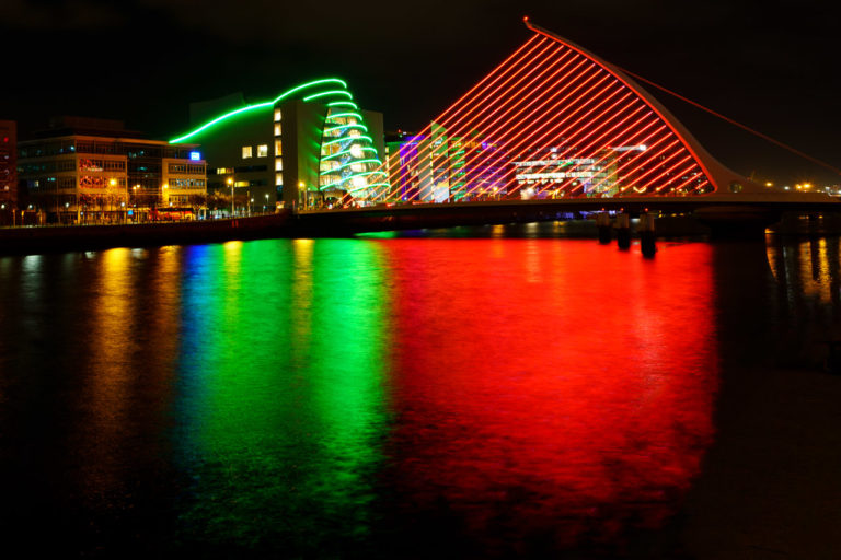 Lights reflect off a bridge and nearby building on the waters of a river at night