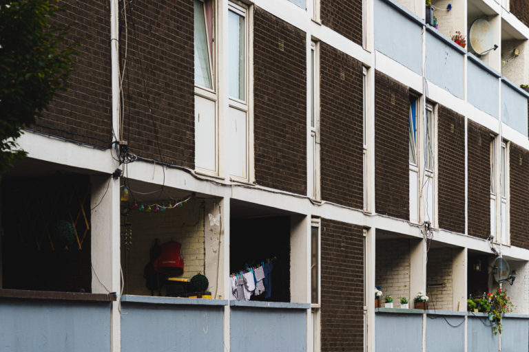 The back of a block of flats. Washing hanging up, flowers decorate the area. A satellite dish pokes out.
