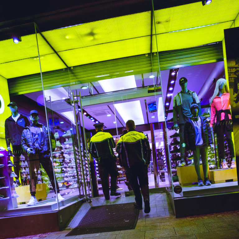 Two Gardai enter a shop. The shop has a bright yellow border matching the colour of the shoulder area of the Gardai uniforms