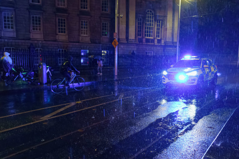 A Garda (police) car driving along a street while rain falls at night. A cyclist is on the rother side of the road and pedestrians walk on the pavement.