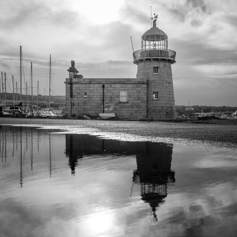 Black and white image of Howth Harbour Lighthouse with a reflection in a puddle of water.