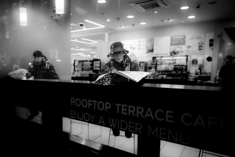 A woman reads a newspaper and a man looks at his coffee in a cafe, through the front window.