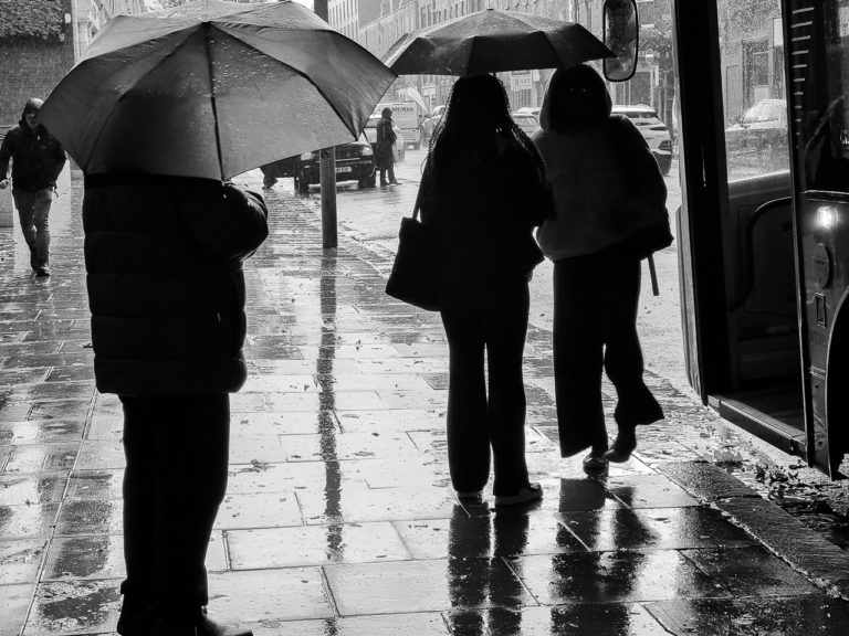 A man sheltering under an umbrella as two young ladies walk off.