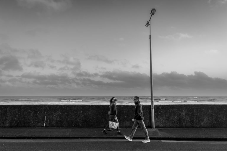 A man walks on the road while two women walk on the pavement in the opposite direction. A wall and lamp post behind them, and the sea is visible over the wall.