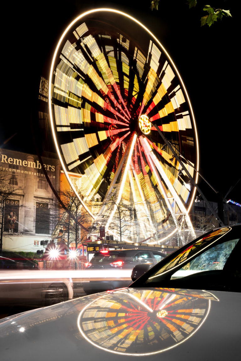 A spinning ferris wheel is reflected in the bonnet of a nearby parked car