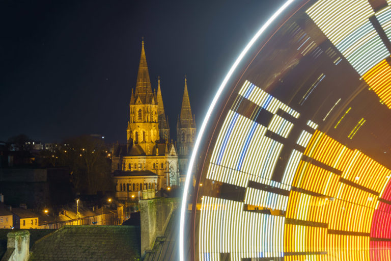 A long exposure photo of a quarter of a Ferris Wheel with Saint Fin Barre's Cathedral in the background