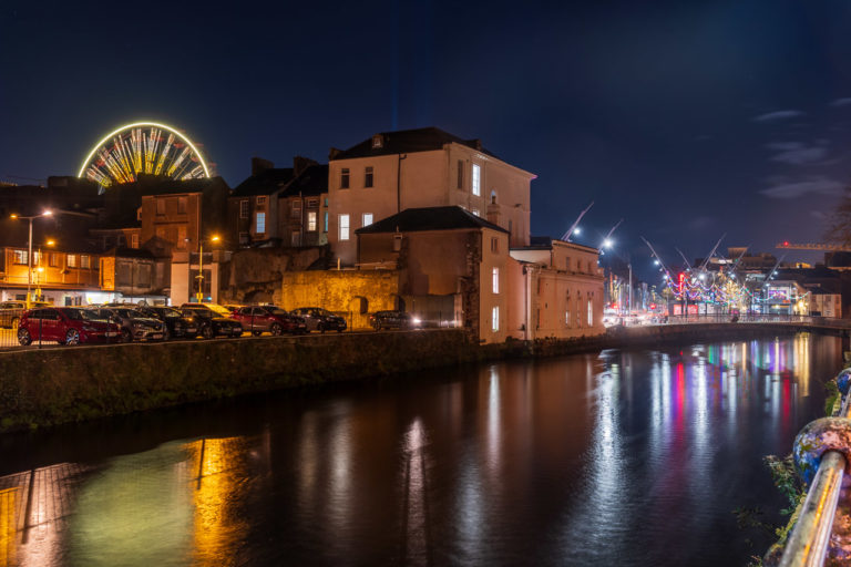 Light reflected in the water of a river at night in a city. Bright Christmas decorations line the bank further down, while the top half of a Ferris Wheel is lit up and seen above local buidlings. A car park stands next to the river.