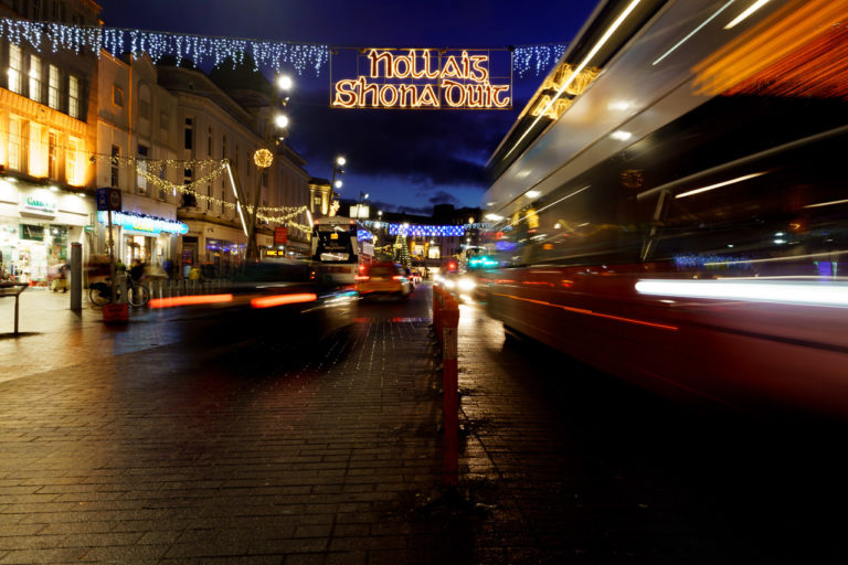 A bus and cars speed along the road as a sign above says "Nollaig Shona Duit".