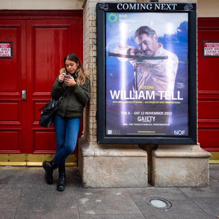 A woman checks her phone while leaning against a wide pillar with a poster advertising the play, William Tell.