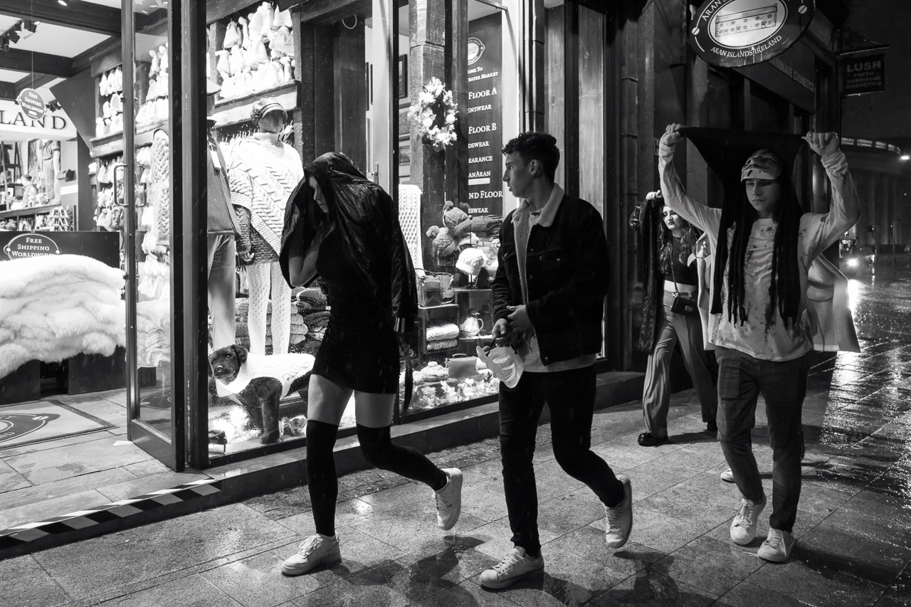 A group walking in the rain, some covering their heads with jackets. One young man looks in a shop window.