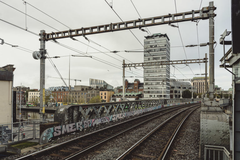 A bridge over the River Liffey with train tracks running over it. Liberty Hall and other buildings are seen in the background.