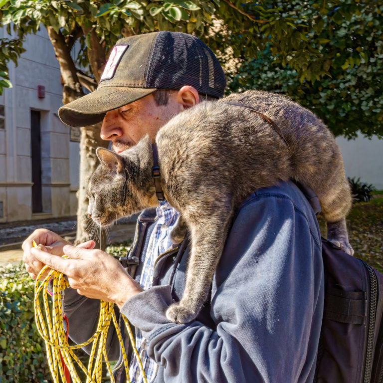 A man puts some kind of paste on his finger while being watched carefully by his cat who is lying over his shoulder!