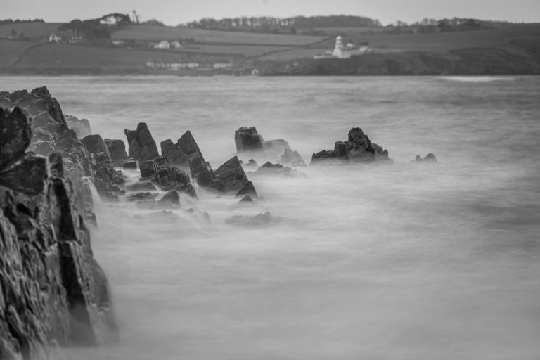 Water crashes on rocks but is turned to mist in a long exposure photo, with a lighthouse in the background.