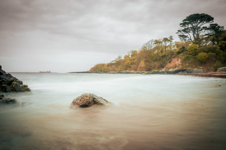 A long exposure photo of the sea smoothes out the waves. A ship sits at anchor on the horizon.