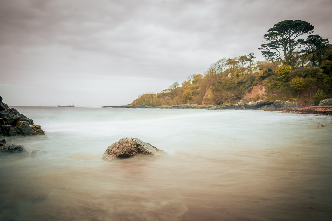 A long exposure photo of the sea smoothes out the waves. A ship sits at anchor on the horizon.