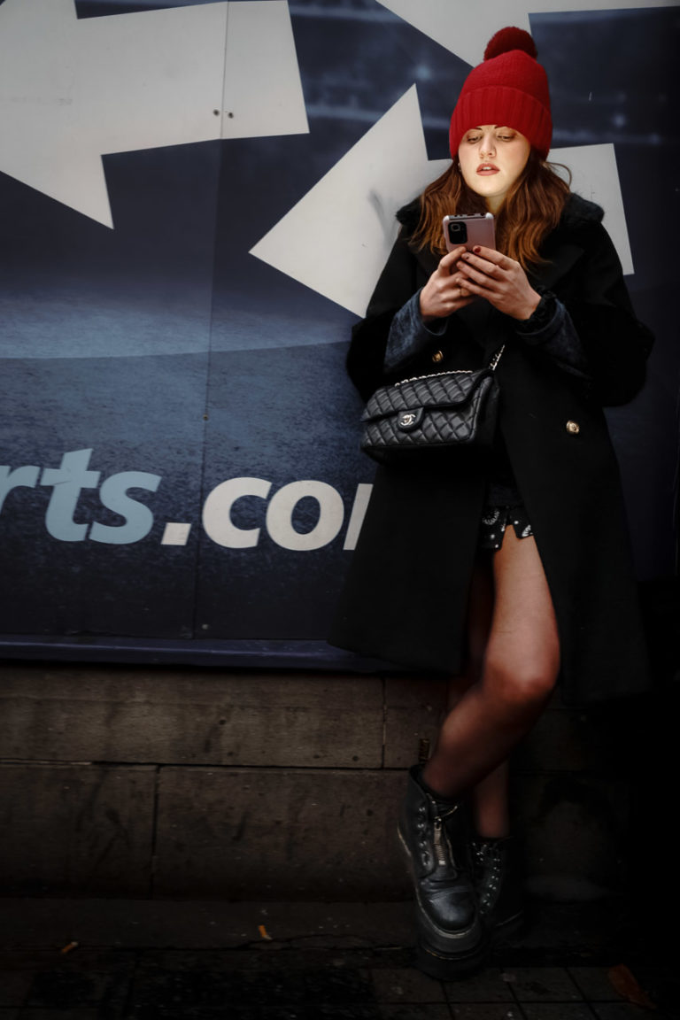 A young woman in a black coat, boots and a red wooly hat checks her phone while leaning against a wall.