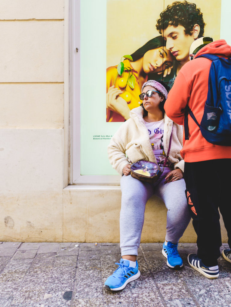 A woman sits on a window sill, while a man (her partner?) stands next to her. Another couple in a photo in the window look on.
