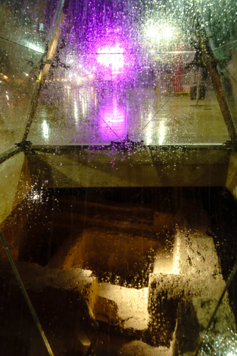 A wide angle photo of the glass pyramid in Malaga. Roman ruins underneath and bright purple LED lights above.