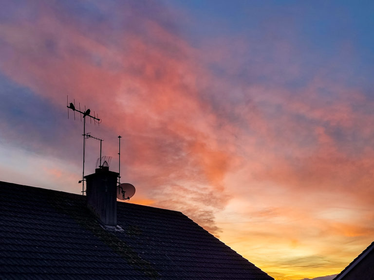 Two birds sitting on a TV ariel on a house, as the sun rises behind them casting lovely colours on the sky.