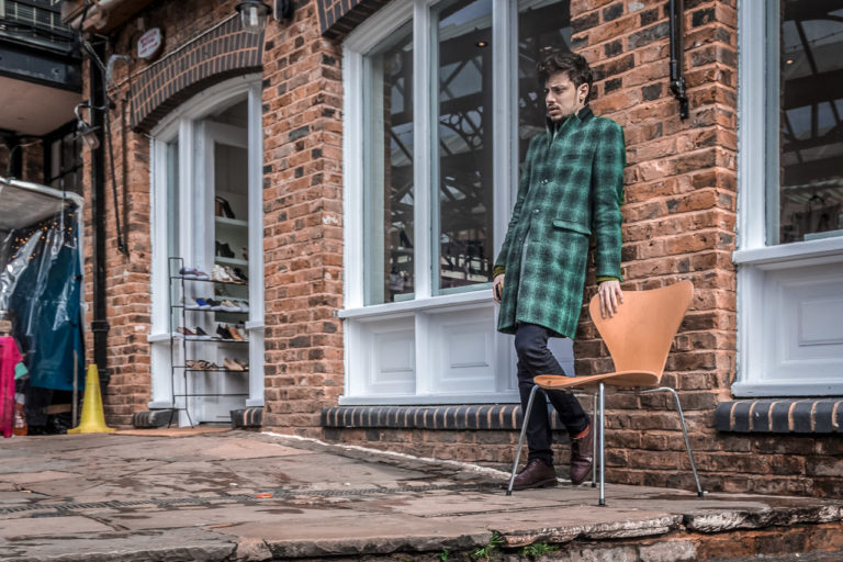 A young man in a green patterned coat leans against a wall outside a shoe shop in Camden Town, London. He looks pensive with a hand on a chair, while clothes hang in the background, adding to the lively atmosphere.