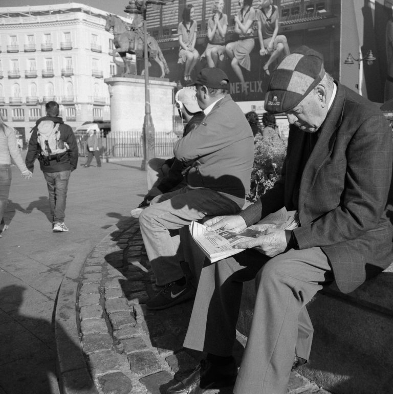 An older man wearing a cap that says "Ireland" on it reads the newspaper. Other men sit near him while a couple walk past. A statue of a horse and rider in the background and a large poster advertising a Netflix show is prominent in the background.