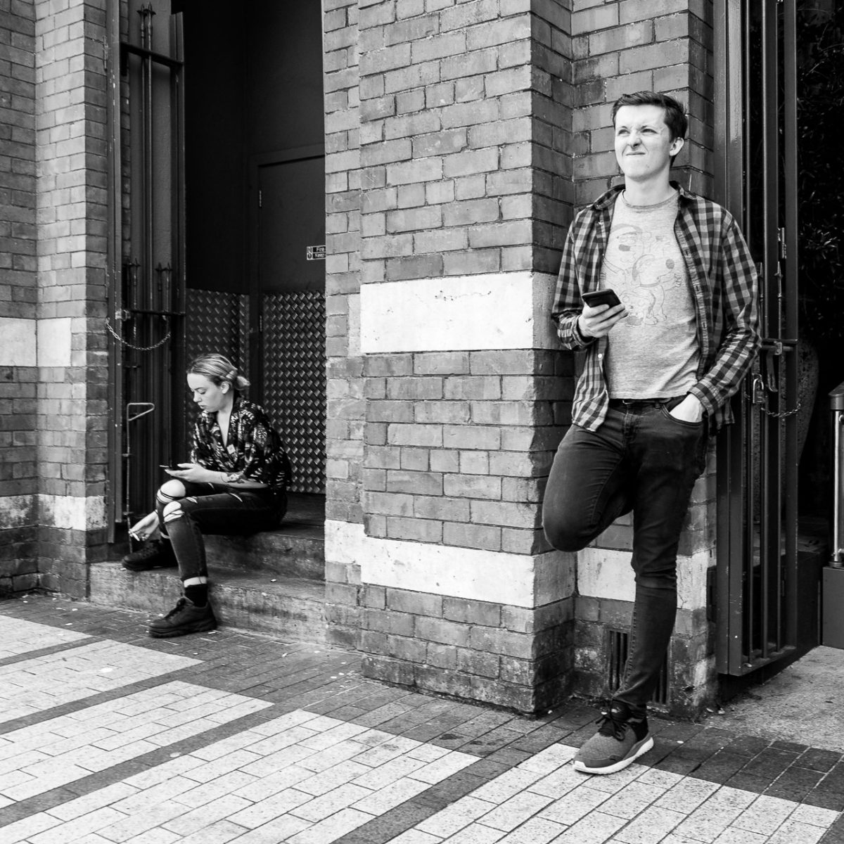 A young man leans against a wall and squints into the bright sky opposite, while a young woman sits on steps nearby checking her phone.