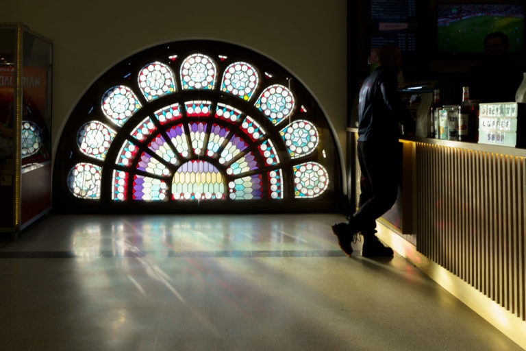 Light streams in through a window illuminating the floor and the bar of a coffee shop. A man leans against the bar, waiting to pay for his purchase.