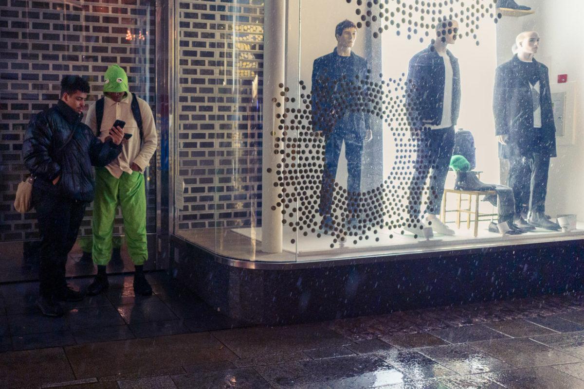 2 young men share a moment, looking at a phone in a doorway, sheltering from the rain. 3 mannequins in a shop window stare out into space.