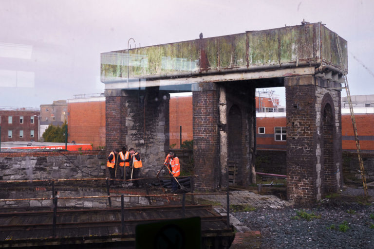 Men repairing the ground near a train track. A large container on brick lined pillars and a wall stand next to them. One person is working while three look at a phone