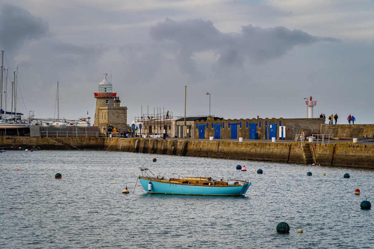 A small boat in the harbour at Howth, Co Dublin. The old and new lighthouses can be seen in the background. People walk along the pier.