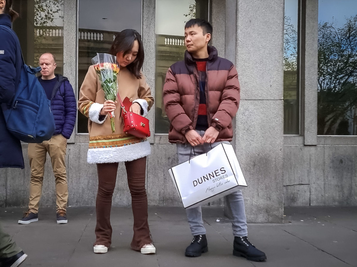 A couple waiting for the bus in Dublin. She has a bunch of flowers and a red handbag she's looking in, he is carrying a Dunnes Stores bag