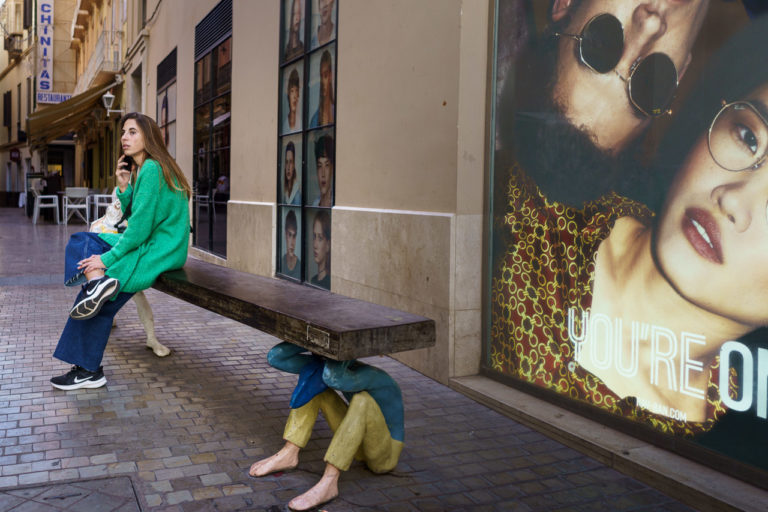 A woman sitting on a bench on the phone. The bench is held up by what looks like a human figure on one end, and barely visible behind her a dog on the other. Two people stare out of a poster, with the text, "You're On"