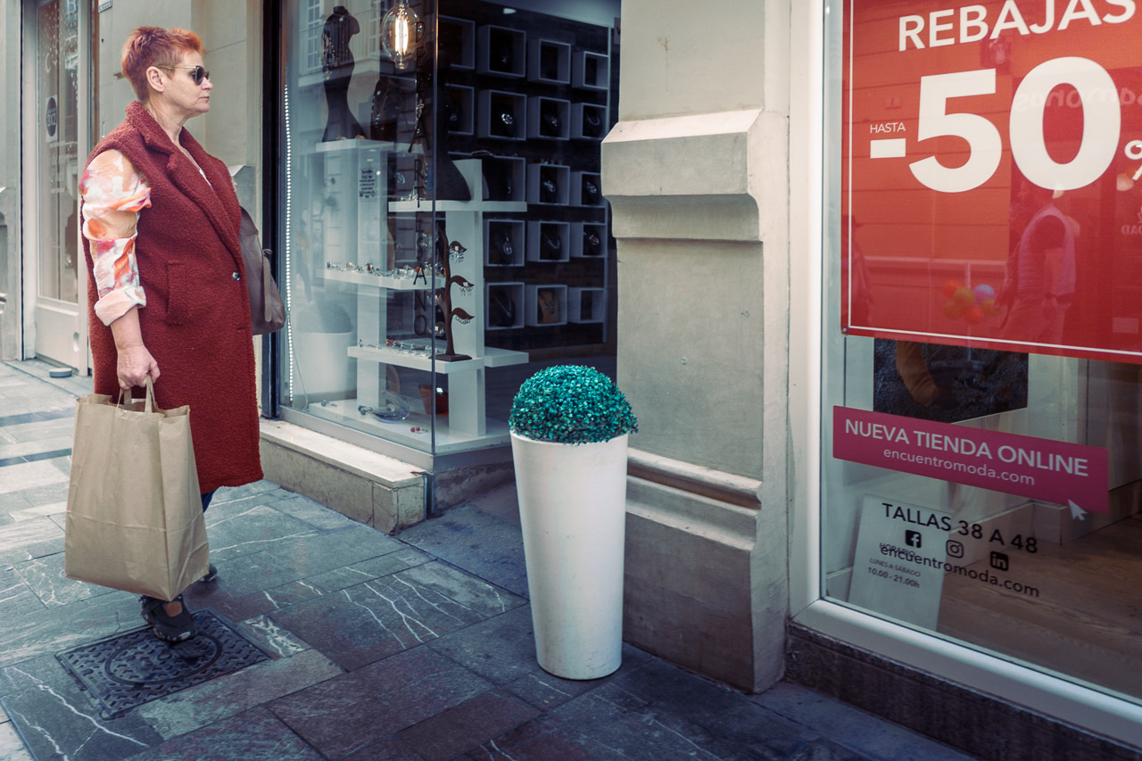 A woman dressed in red looks in the door of a shop. In the window can be seen a sign saying, "REBAJAS HASTA - 50%"