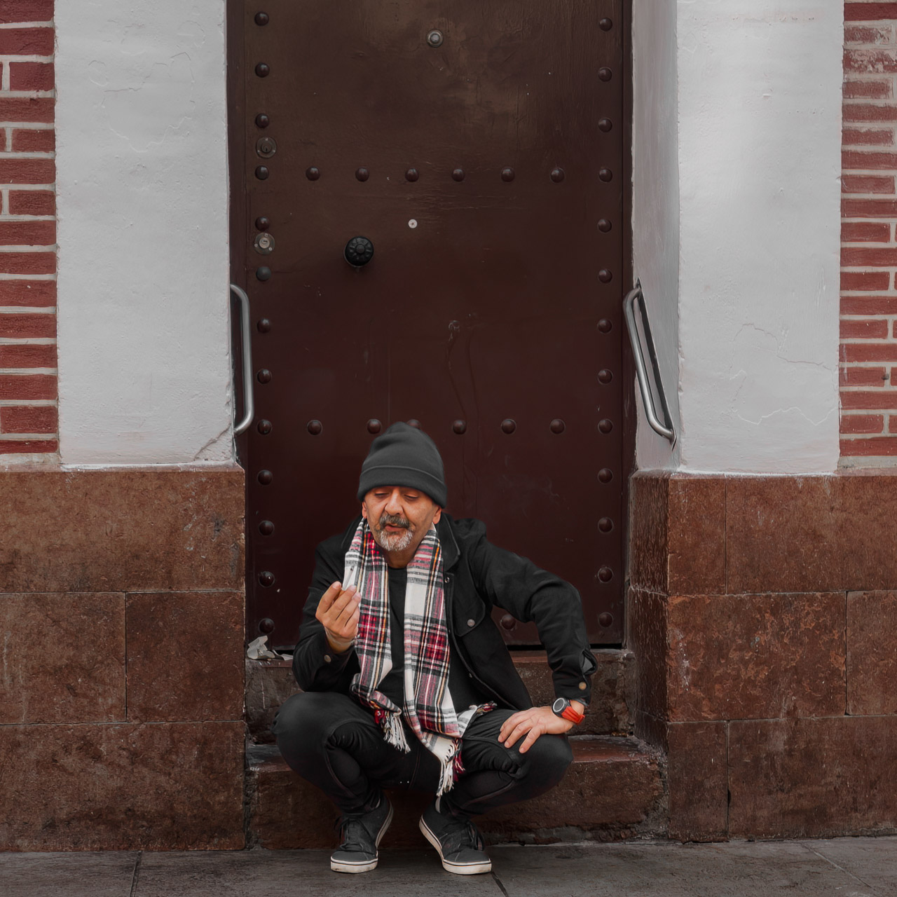 A man contemplates his cigarette while sitting on the steps of a building. He's wearing a woolly hat and a scarf as it's late December in Malaga.