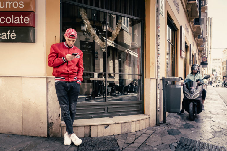 Two young men, both on their phones. One leaning against the wall, the other a delivery rider on his motorbike.