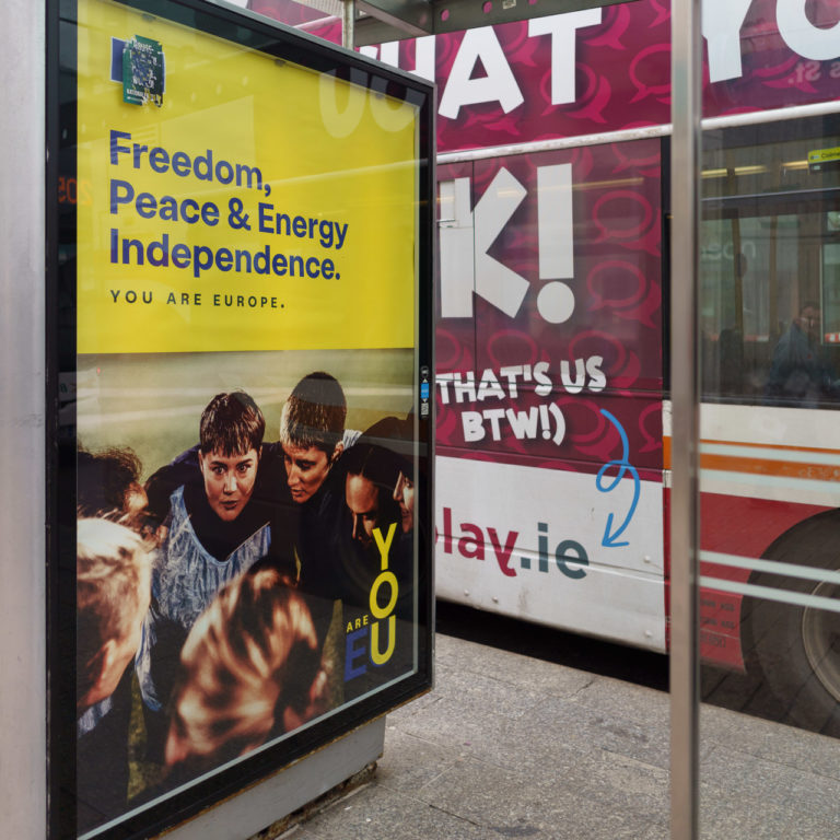 A poster on a bus shelter proclaims "Freedom, Peace & Energy Indepence. YOU ARE EUROPE" A group of young people are huddled together next to the text reading "YOU ARE EU" An advert on the bus at the bus stop says "THAT@S US BTW!" with an arrow pointing down to the end of a URL.